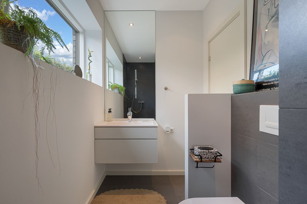 Modernes Badezimmer mit großem Spiegel, Hängewaschbecken, Dusche und Pflanzen am Fenster.