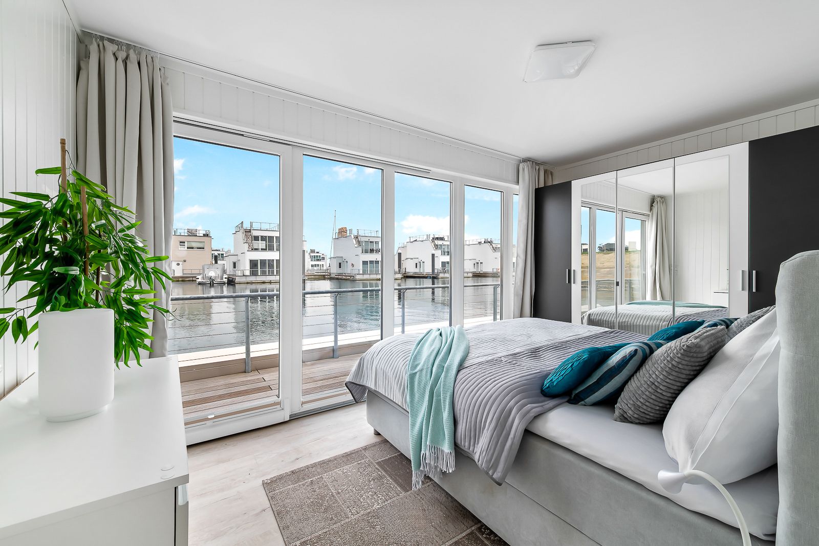 Helles Schlafzimmer mit großem Bett, Spiegelschrank und Blick auf das Wasser durch große Fenster.