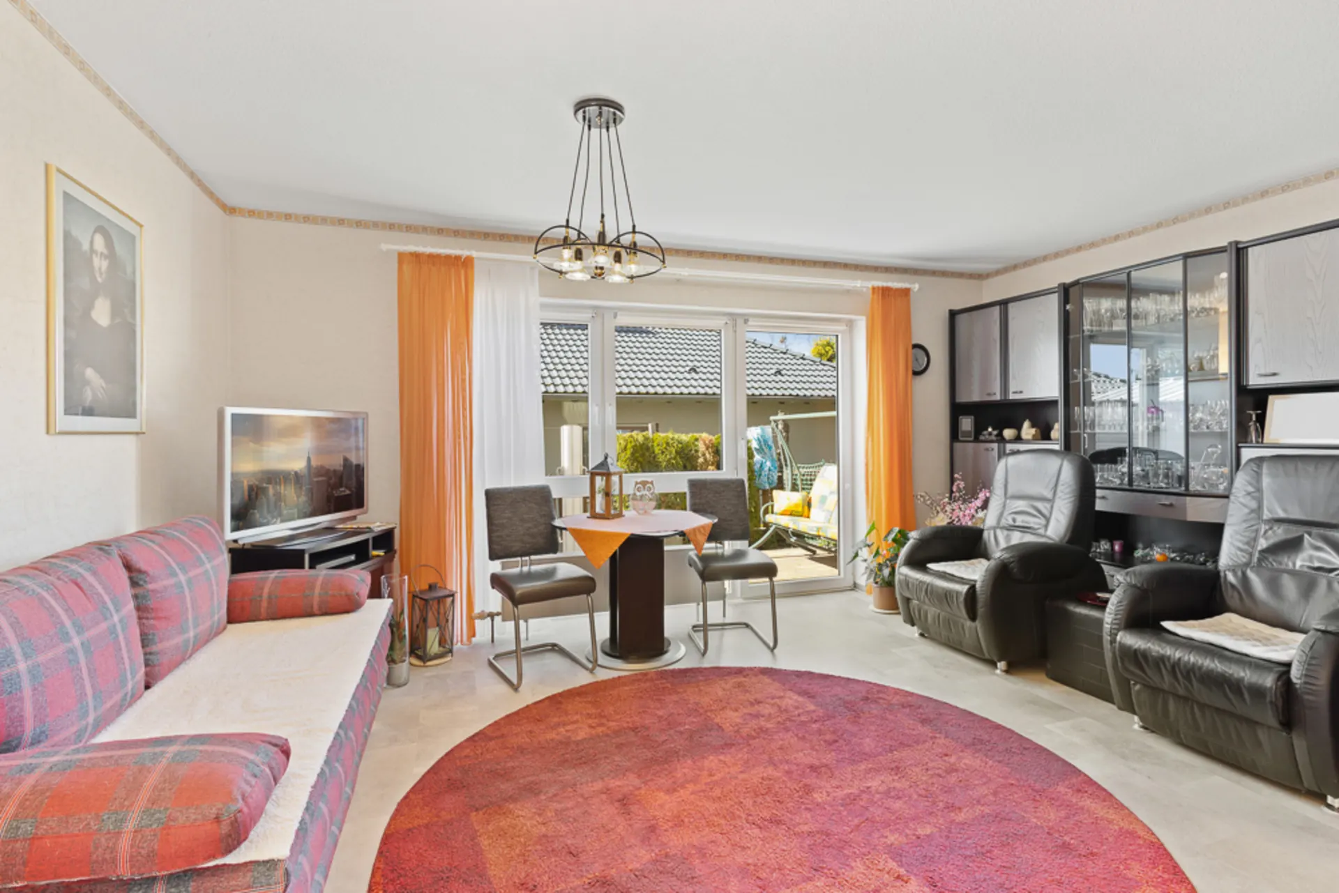 Wohnzimmer mit Sofa, zwei Sesseln, Esstisch, Glasvitrine und Zugang zur Terrasse.