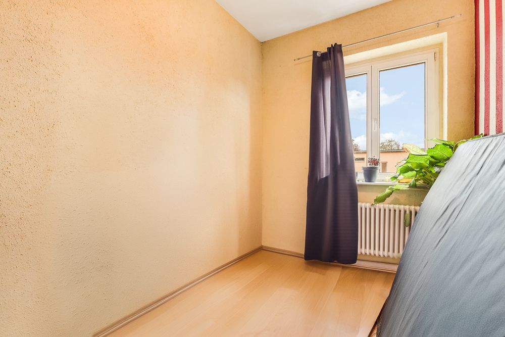 Kleiner Raum mit hellem Holzboden, Fenster, schwarzem Vorhang und zwei Pflanzen auf der Fensterbank.