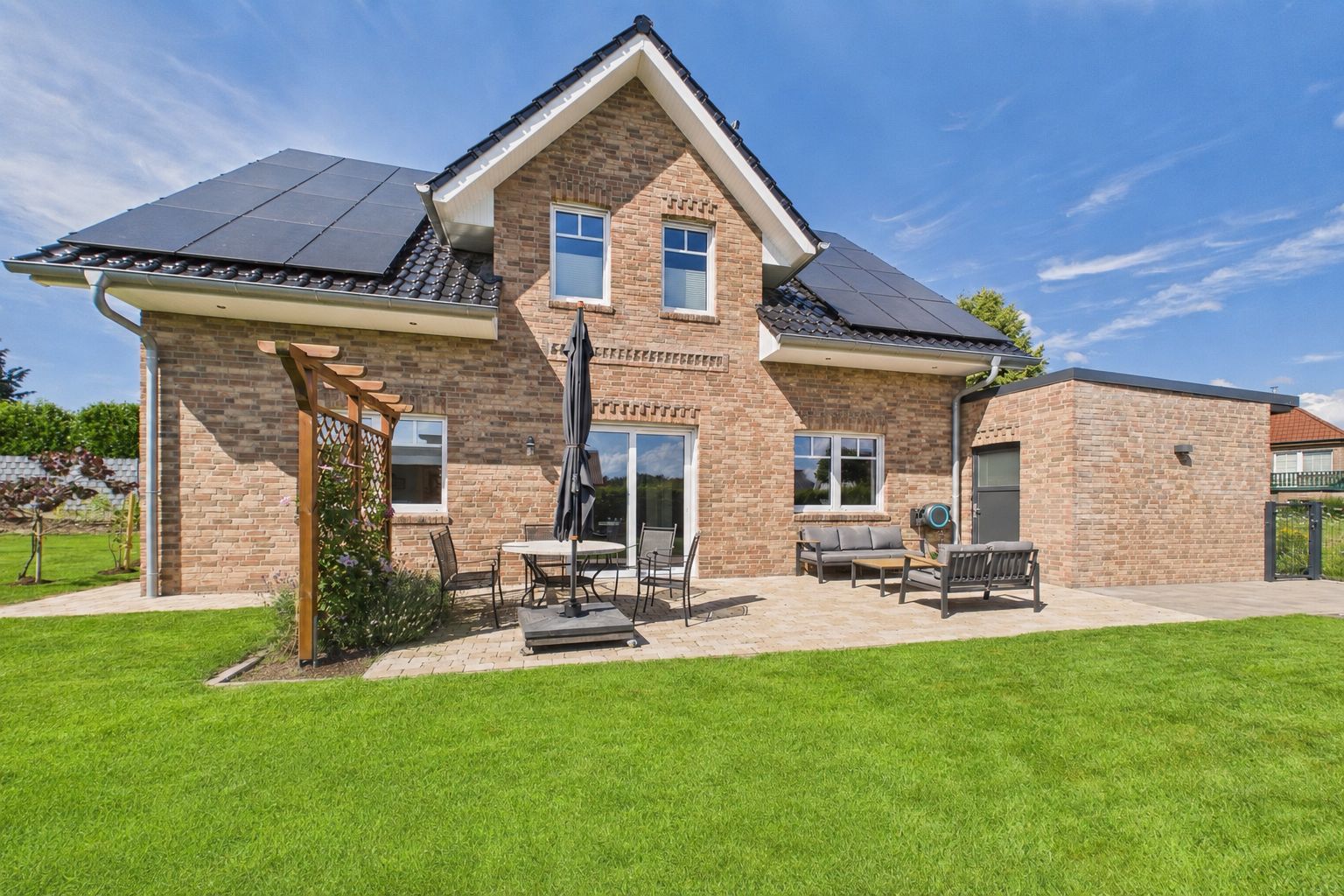 Backsteinhaus mit Solarpanelen, Terrasse mit Sitzmöbeln und grünem Rasen im Vordergrund.