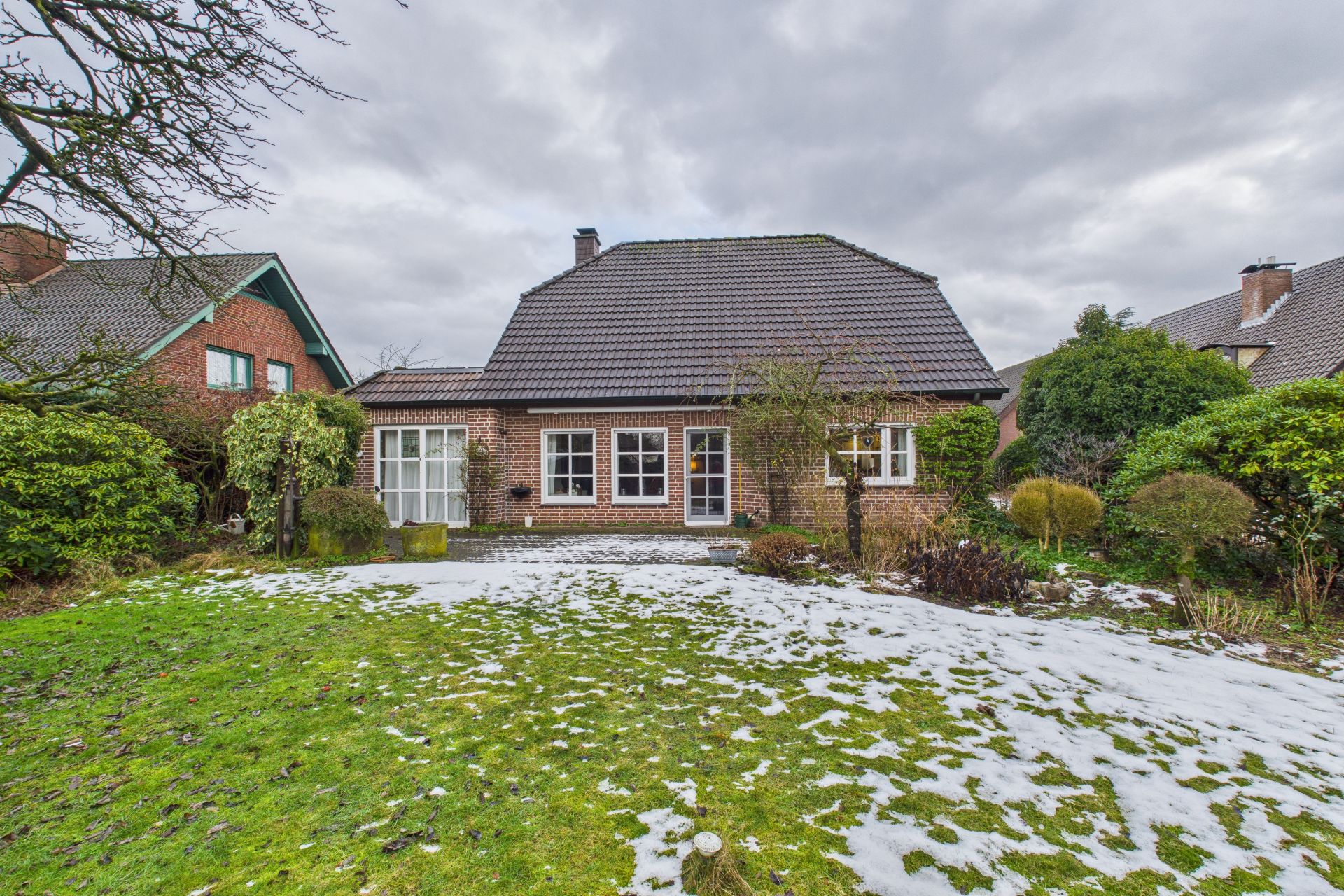 Backsteinwohnhaus mit dunklem Dach, Garten und etwas Schnee auf dem Rasen.