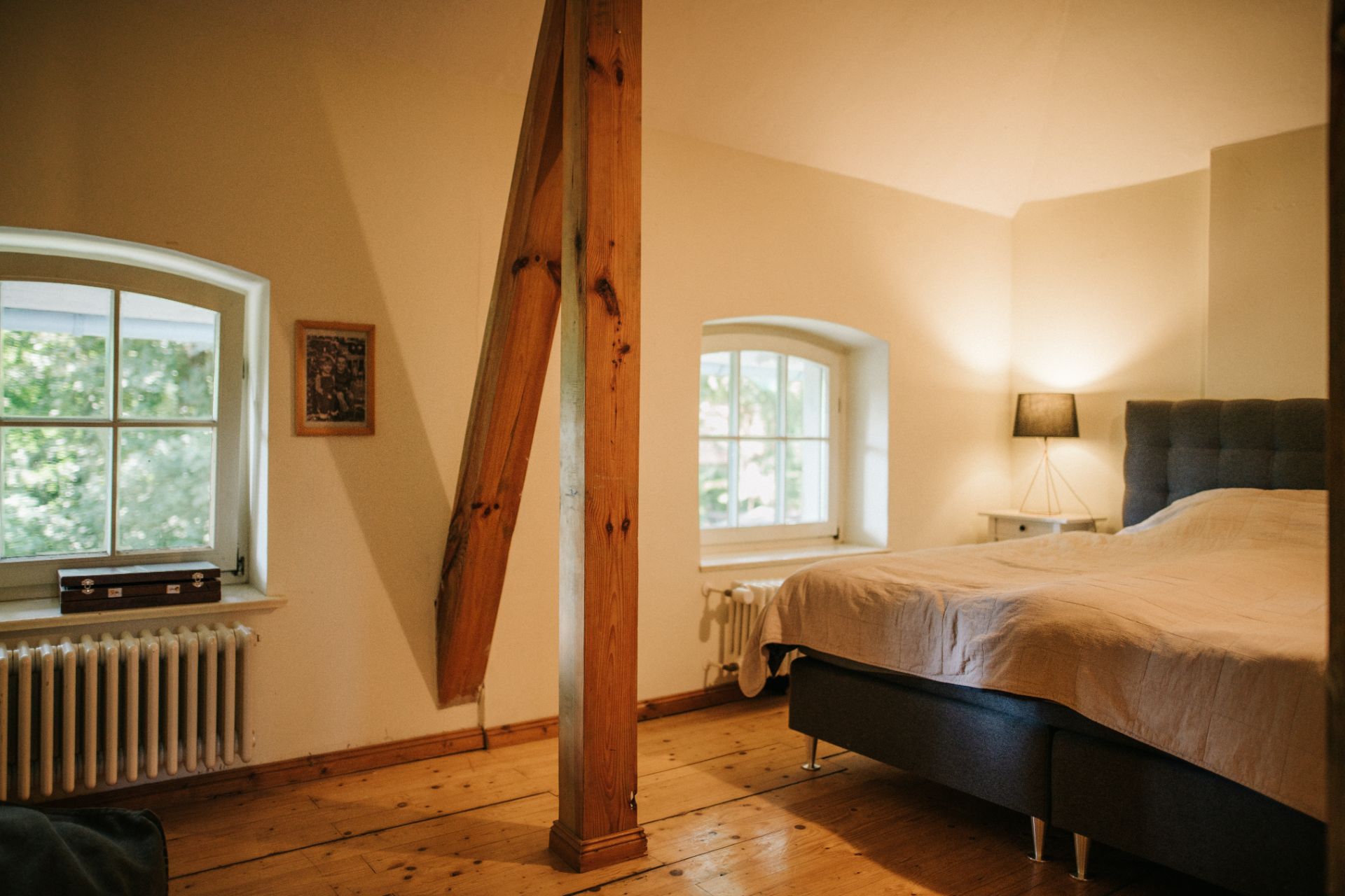 Schlafzimmer mit Holzboden, Holzbalken, zwei Fenstern, Bett und Nachttischlampe.