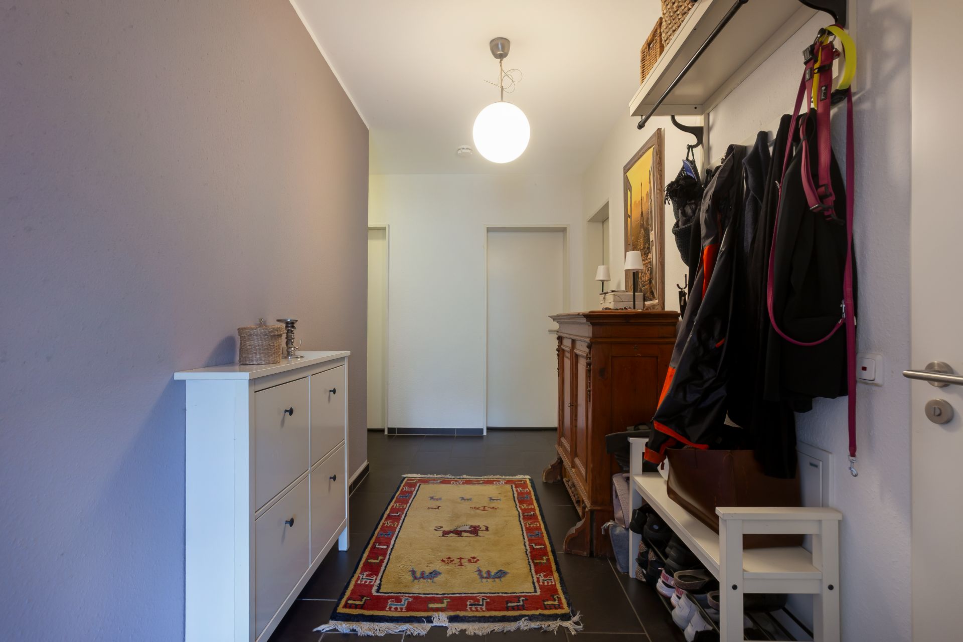 Flur mit Teppich, Schuhschrank, Garderobe und Holzmöbel, helle Wände, Deckenlampe.