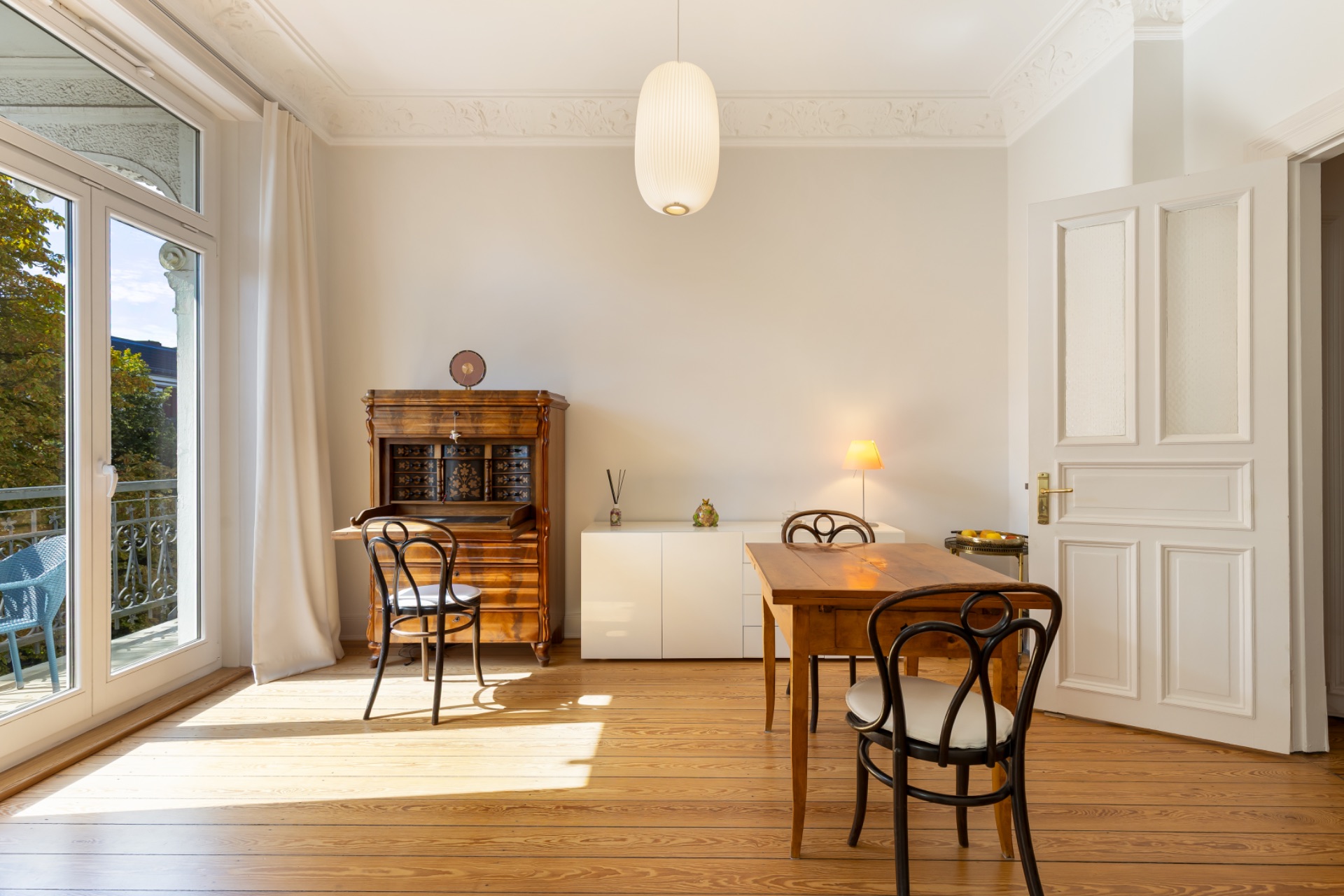 Helles Zimmer mit Holzboden, Tisch, Stühlen, Sekretär und Zugang zu einem Balkon.