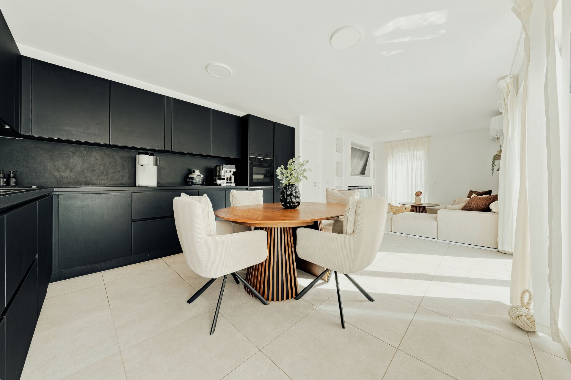 Moderne Küche mit dunklen Schränken, rundem Holztisch und vier Stühlen. Helles Wohnzimmer im Hintergrund.