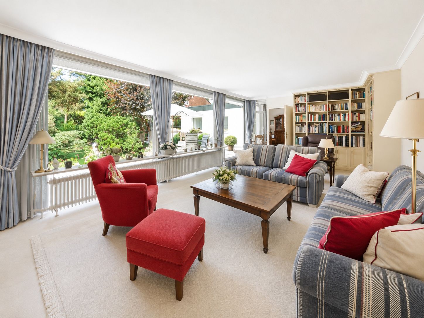 Helles Wohnzimmer mit großen Fenstern, blauen Sofas, roten Sesseln und Bücherregal.