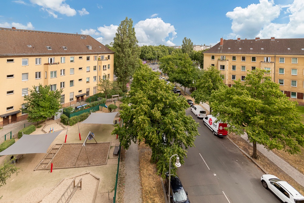 Blick auf Wohnhäuser, Bäume, Straße und einen Spielplatz mit Sandkästen und Schaukeln.