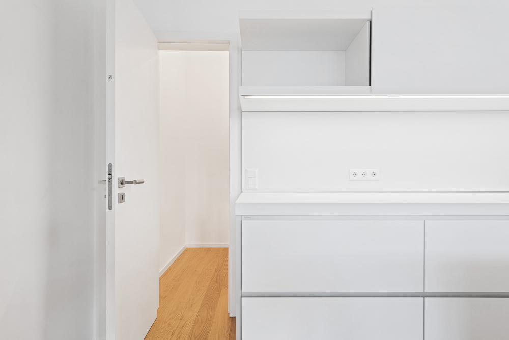 Offene weiße Tür führt zu einem Flur mit hellem Holzfußboden, moderne weiße Möbel im Vordergrund.