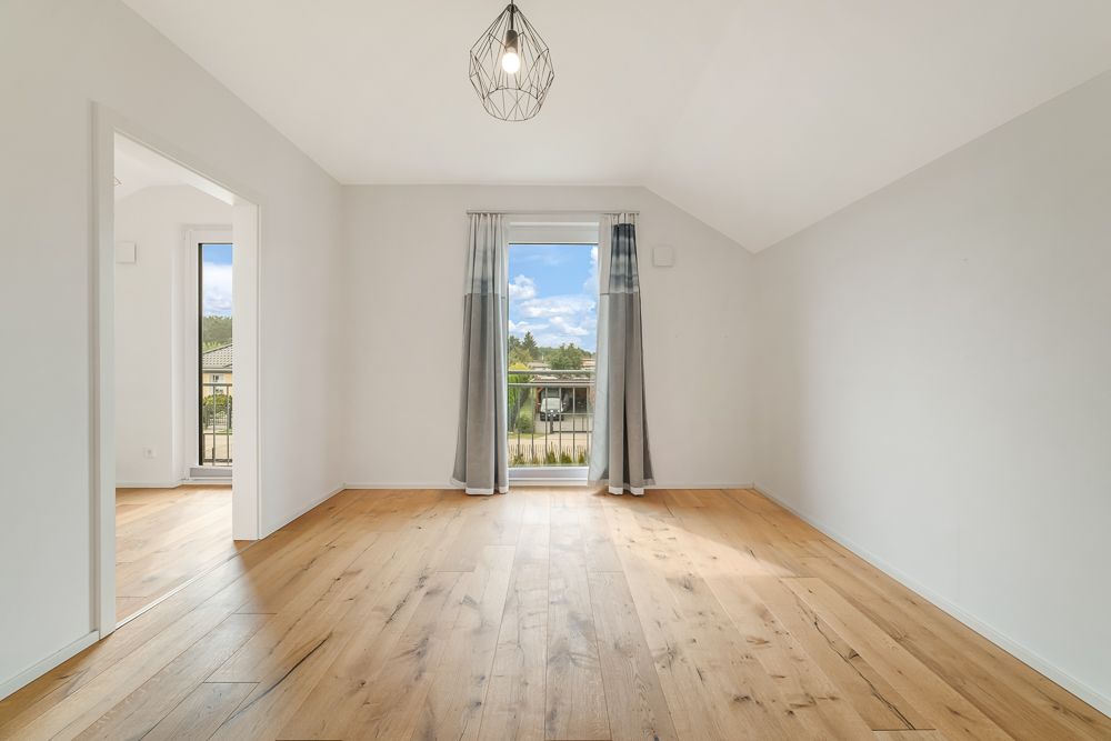 Heller, leerer Raum mit Holzfußboden, Fensterfront mit Vorhängen und Deckenlampe.