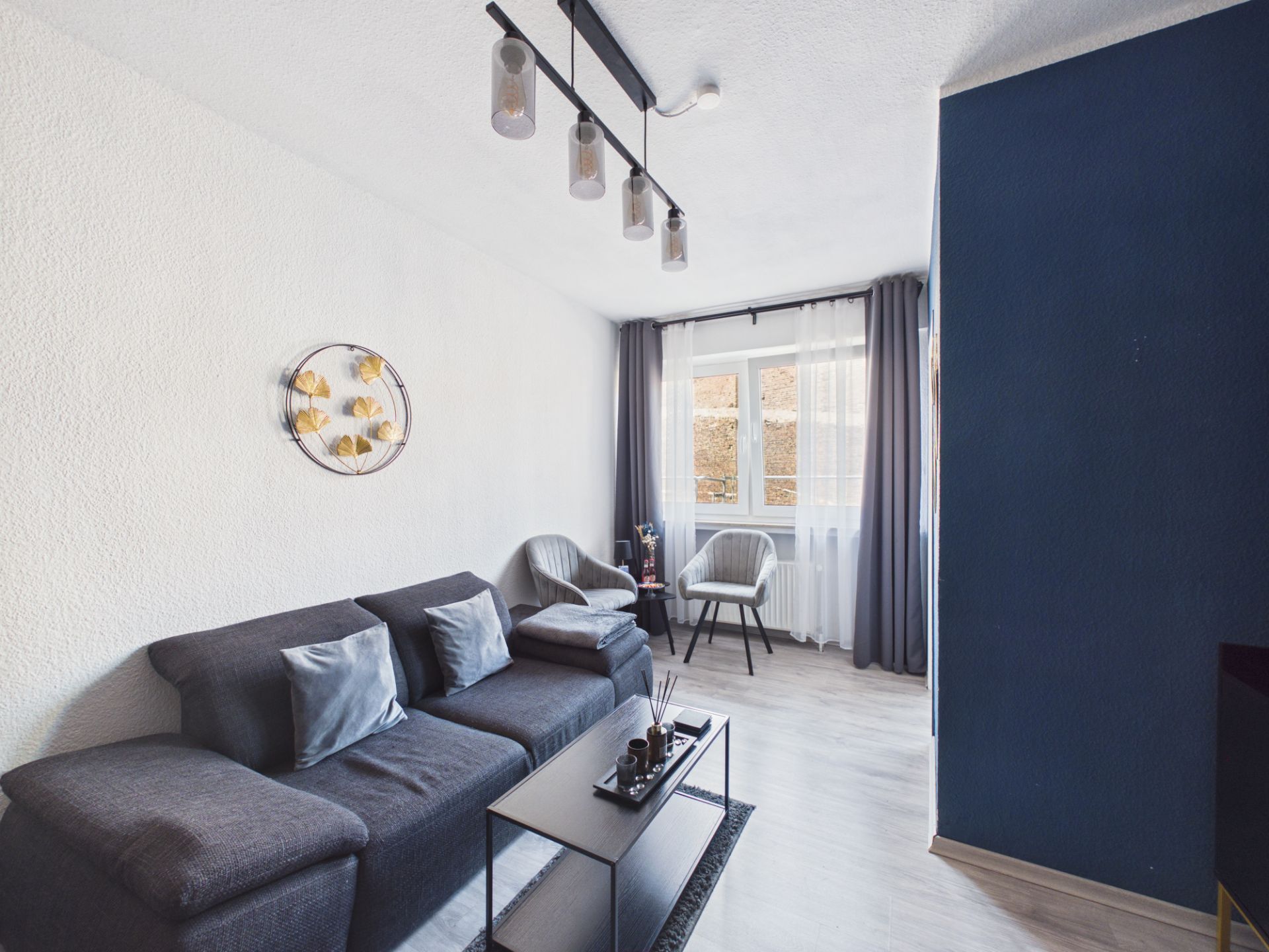 Modernes Wohnzimmer mit dunkelgrauem Sofa, Couchtisch und zwei Stühlen vor einem Fenster mit Vorhängen.