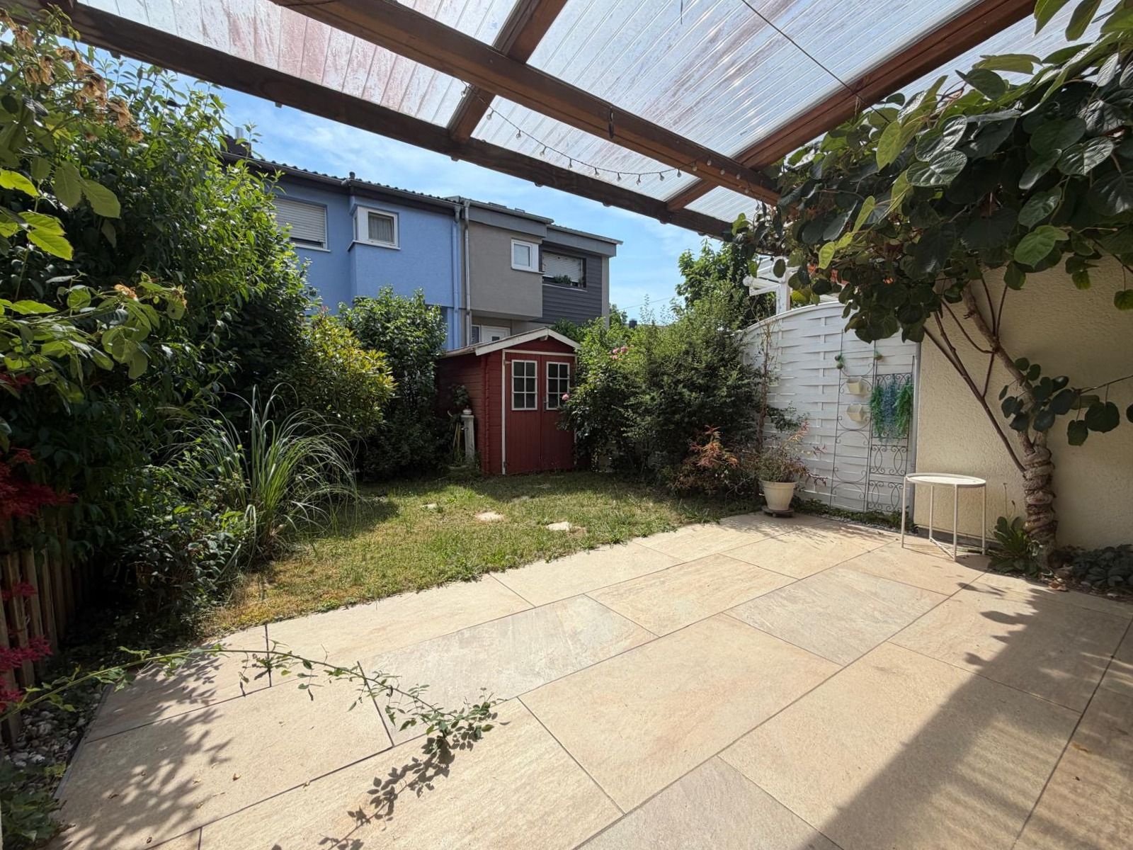 Überdachte Terrasse mit Blick auf einen kleinen Garten und ein rotes Gartenhaus.
