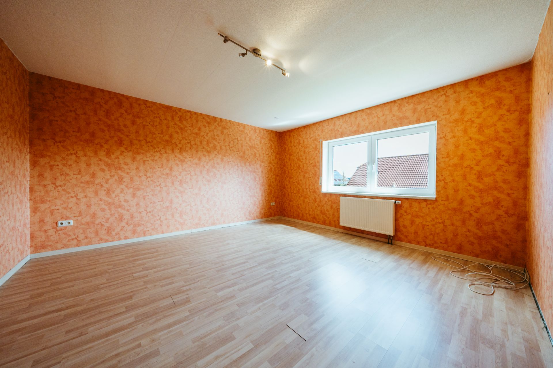 Leeres Zimmer mit orangefarbenen Wänden, großem Fenster und hellem Laminatboden.
