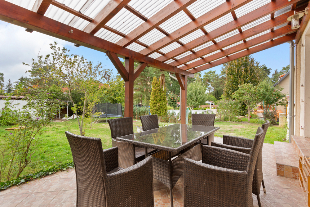 Überdachte Terrasse mit Glastisch und sechs Gartenstühlen, Blick auf grünen Garten.