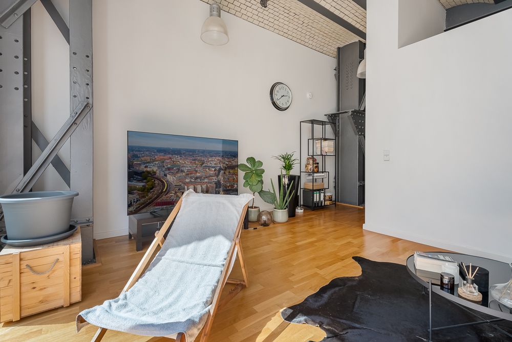 Helles Wohnzimmer mit Liegestuhl, Fernseher, Pflanze, Regal und Wanduhr auf Holzfußboden.