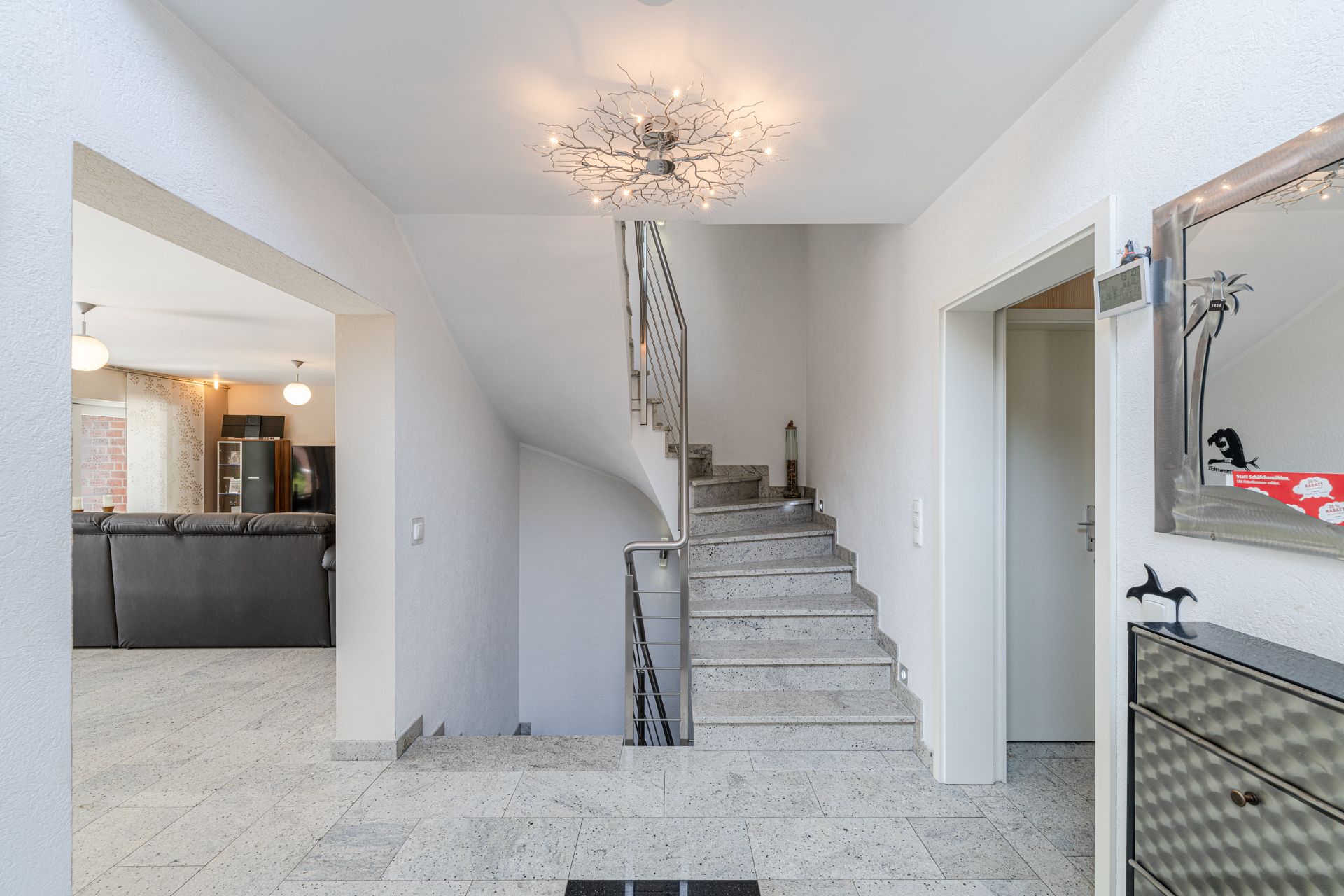Heller Flur mit Steintreppe, modernem Licht und Zugang zu einem Wohnzimmer mit grauem Sofa.