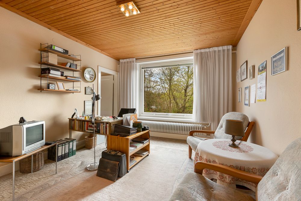 Wohnzimmer mit Sofa, Tisch, Regal, Fernseher und großem Fenster mit Blick ins Grüne.