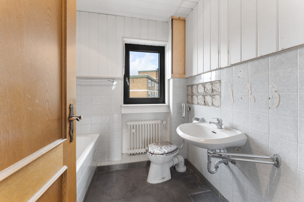 Kleines Badezimmer mit Waschbecken, Toilette, Badewanne und Fenster, sichtbare Gebrauchsspuren an den Fliesen.