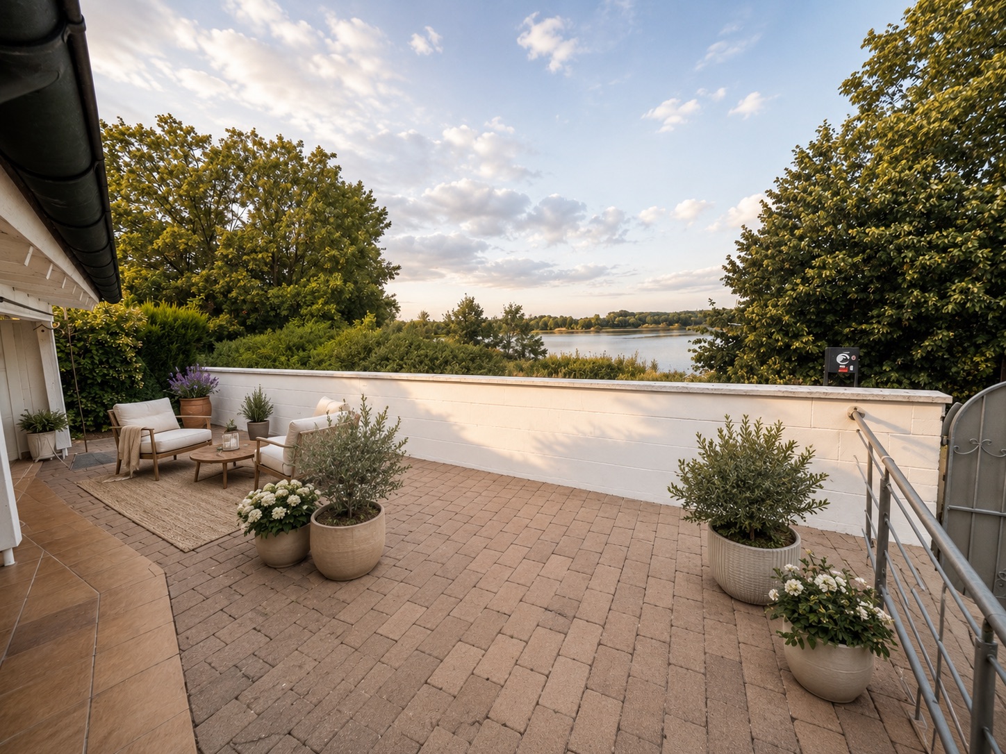 Große Terrasse mit Sitzmöbeln, Pflanzen in Töpfen und Blick auf einen See, umgeben von Bäumen.