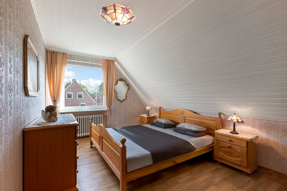 Schlafzimmer mit Holzbett, Nachttischen, Kommode, Fenster und Spiegel an der Wand.