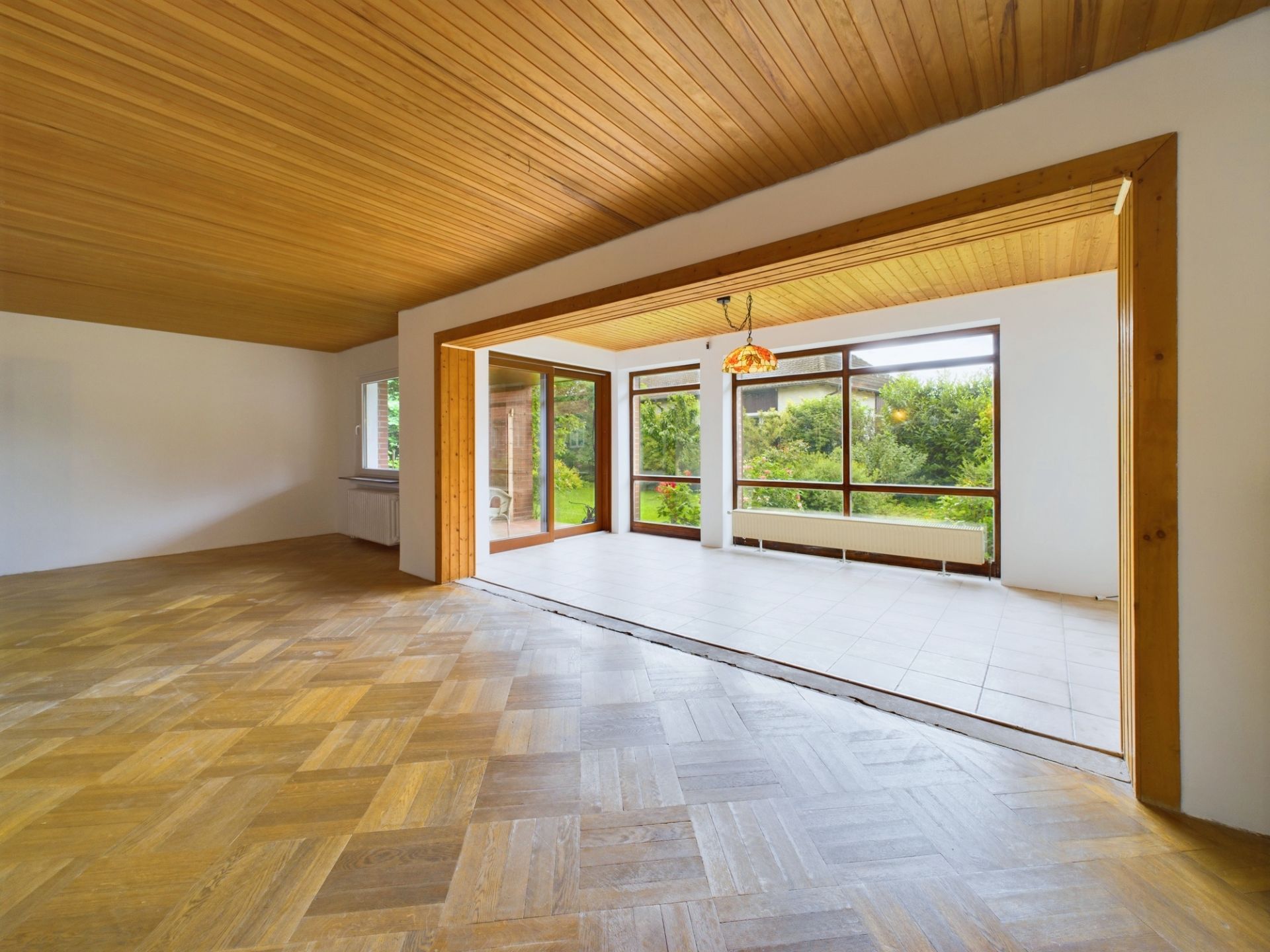 Großer, leerer Wohnraum mit Holzboden und Decke, angrenzender Wintergarten mit Fliesen und großen Fenstern.