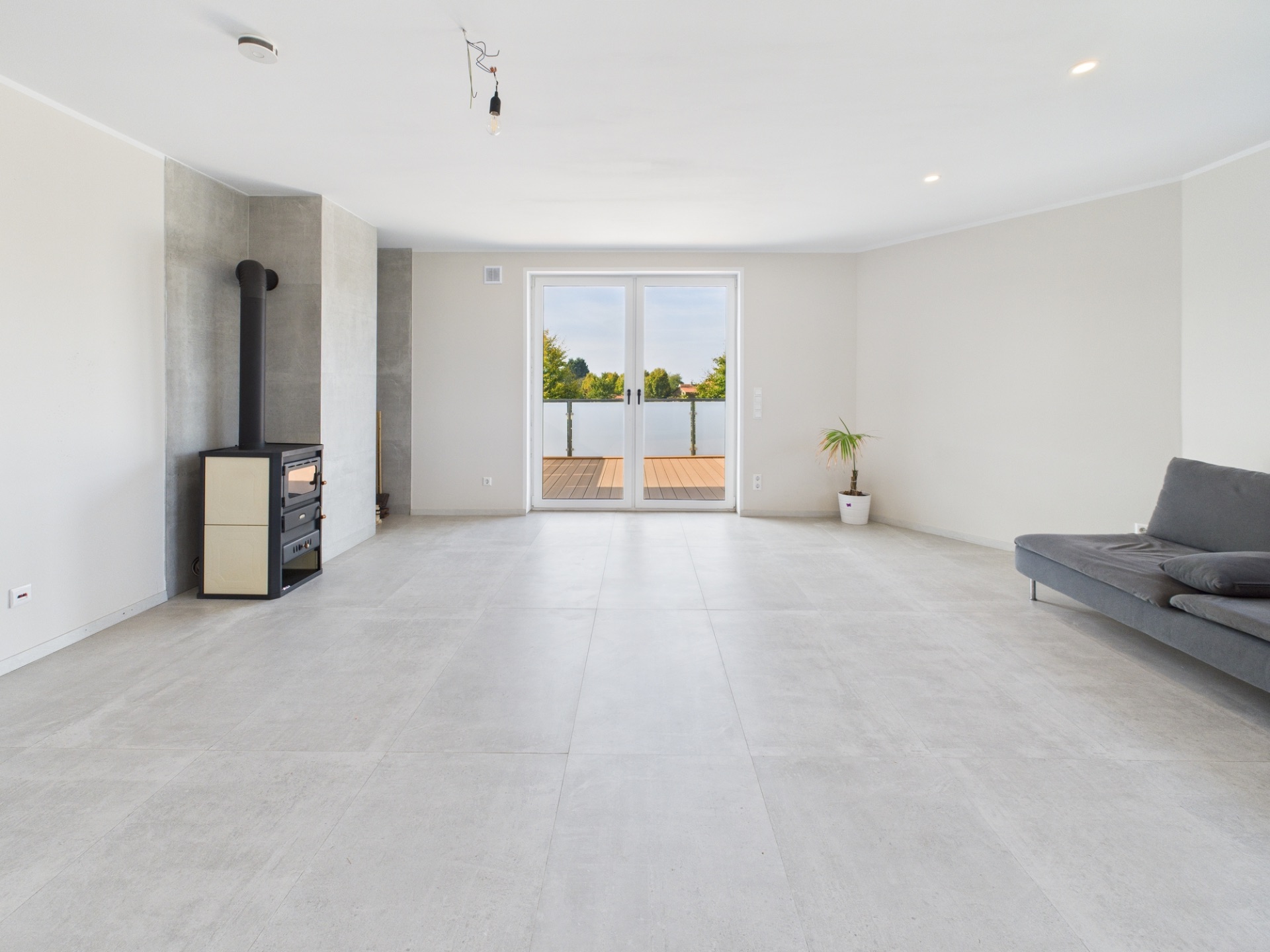 Helles Wohnzimmer mit Kaminofen, Sofa, Topfpflanze und großen Fenstertüren zur Terrasse.