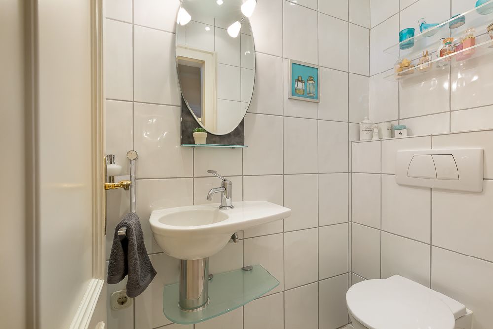 Kleines, modernes Bad mit Waschbecken, Spiegel, WC und Regal auf weiß gefliester Wand.