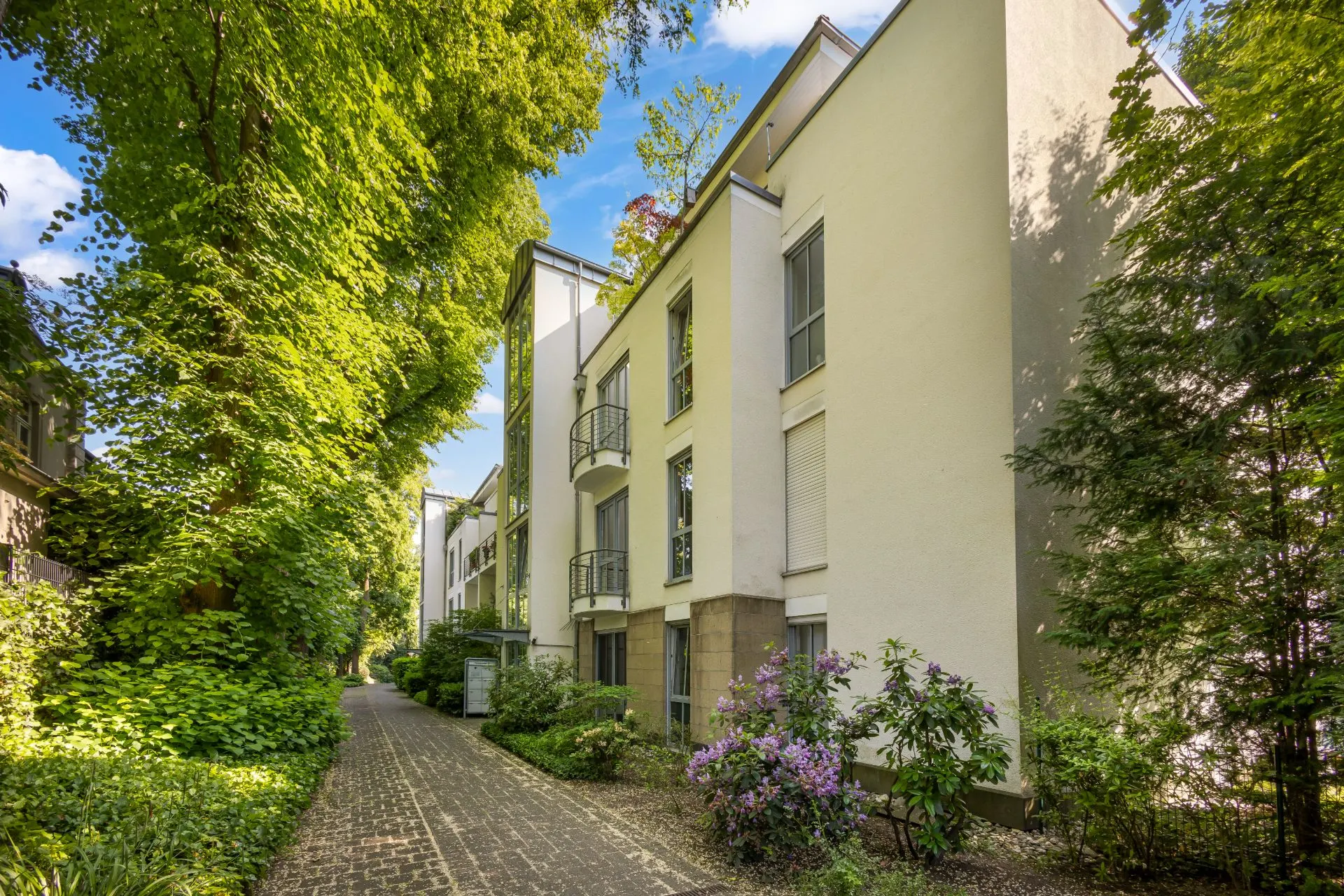 Helles Wohngebäude mit Balkonen, umgeben von Bäumen und Garten, gepflasterter Weg führt am Haus entlang.
