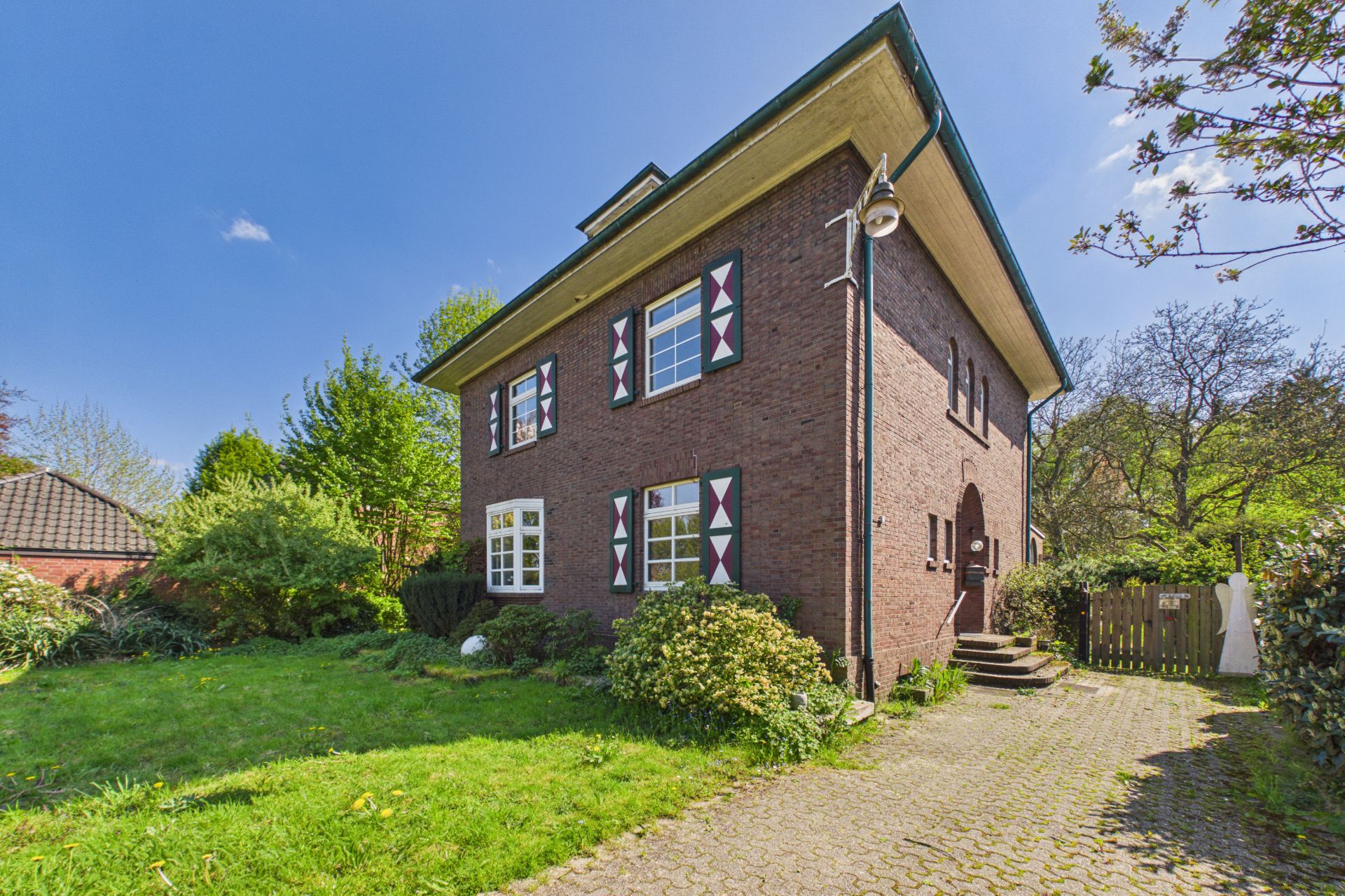 Backsteinhaus mit grünem Garten, roten Fensterläden und gepflastertem Weg vor dem Eingang.