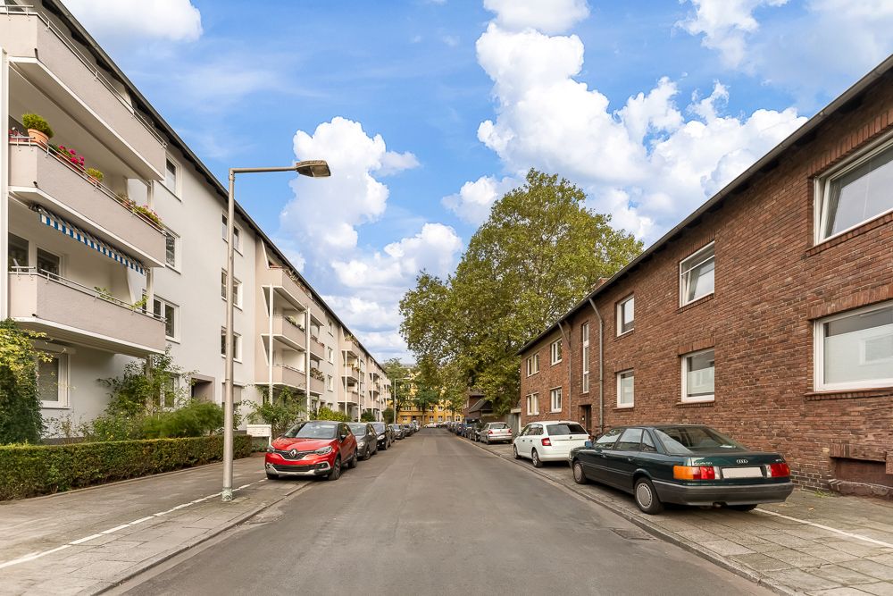 Wohnstraße mit parkenden Autos, Wohnhäusern und Bäumen bei Tag.