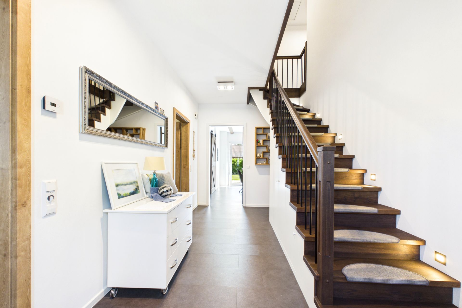 Heller Flur mit dunkler Holztreppe, weißer Kommode und Spiegel an der Wand.