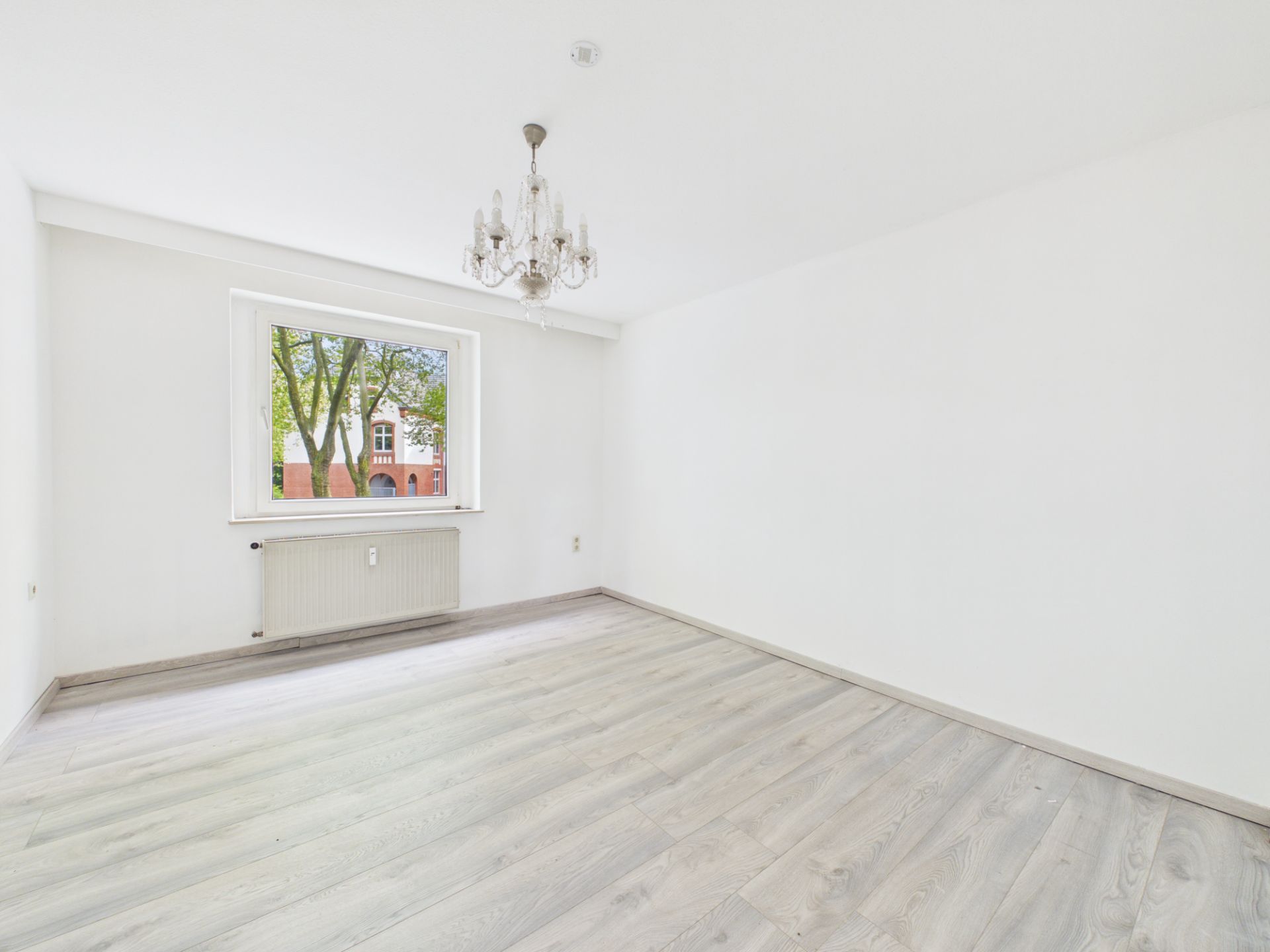 Heller, leerer Raum mit Kronleuchter, großem Fenster und hellem Holzfußboden.