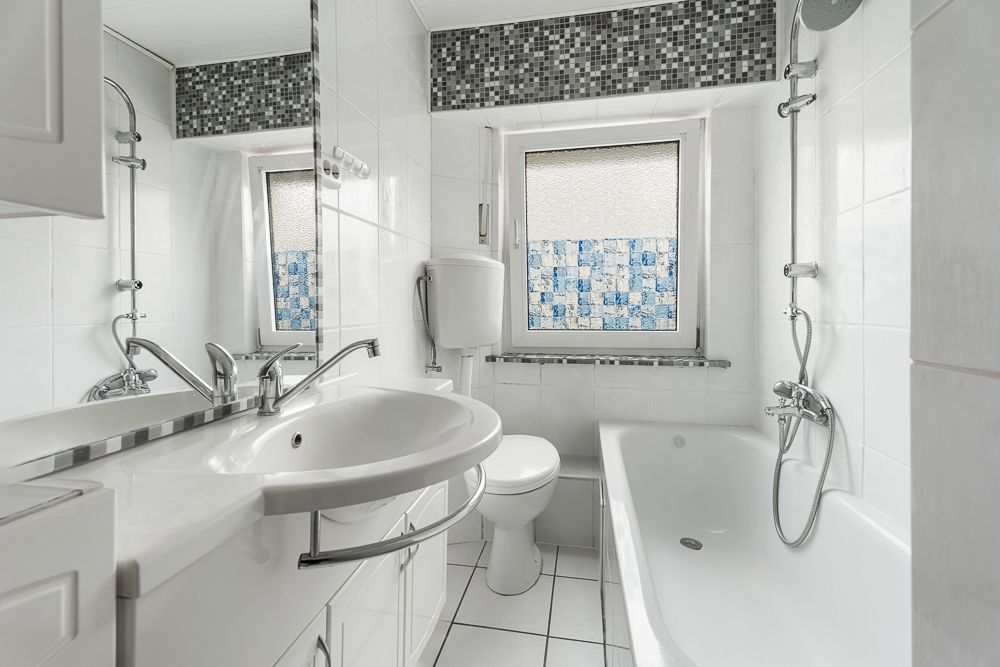 Helles Bad mit Waschbecken, Toilette, Badewanne und Fenster, weiß gefliest.