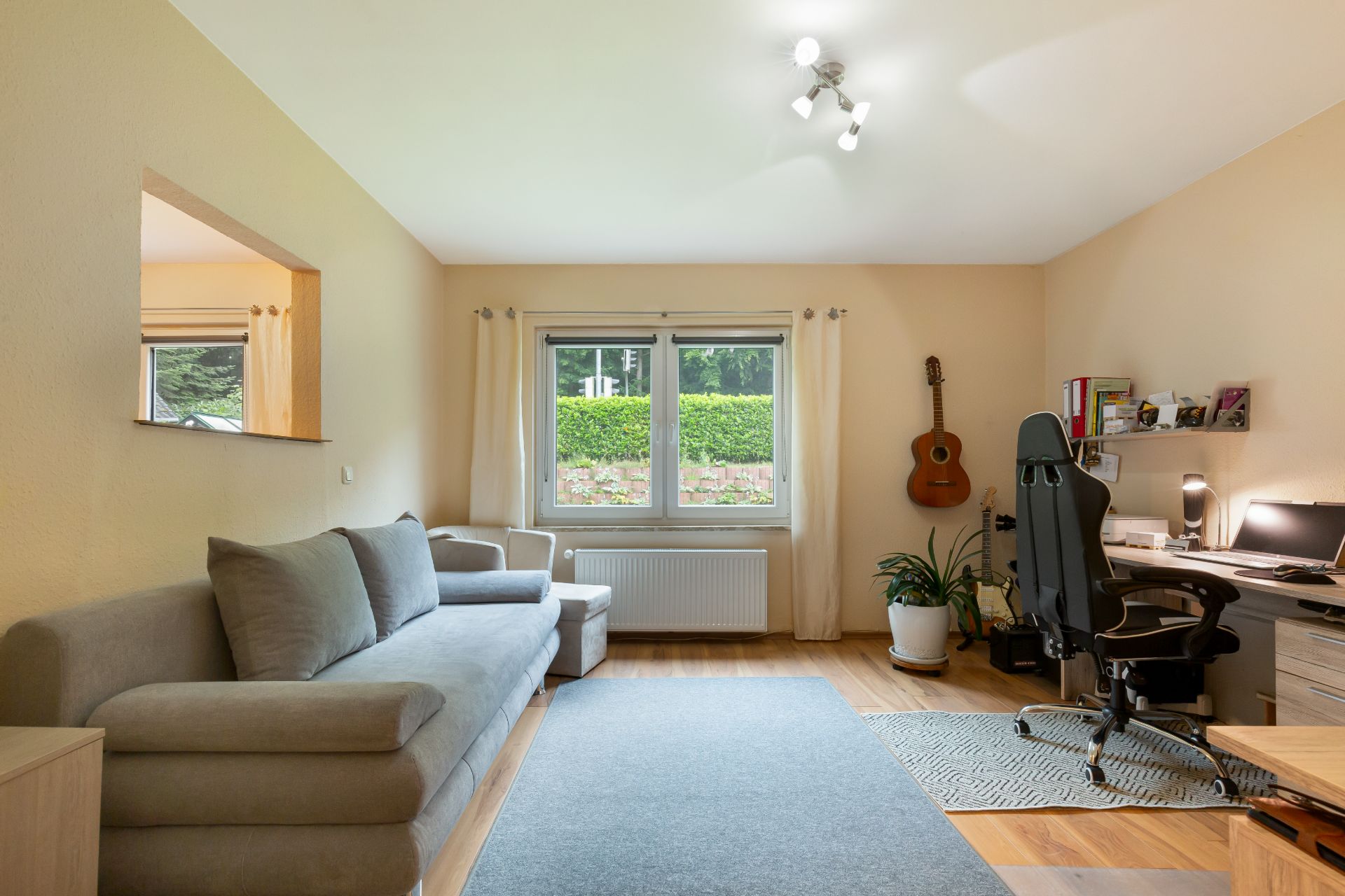 Wohnzimmer mit grauem Sofa, Schreibtisch, Gitarre an der Wand und Fenster mit Blick ins Grüne.