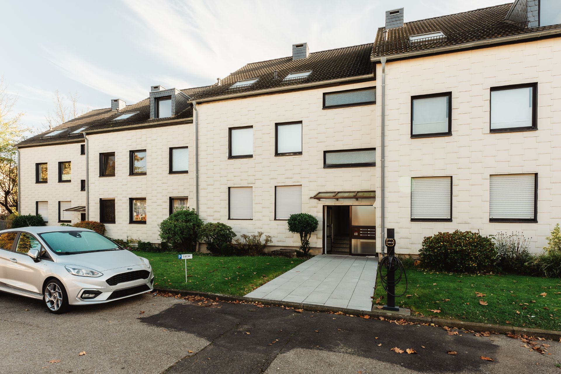 Mehrfamilienhaus mit weißer Fassade, Vorgarten und geparktem Auto davor.