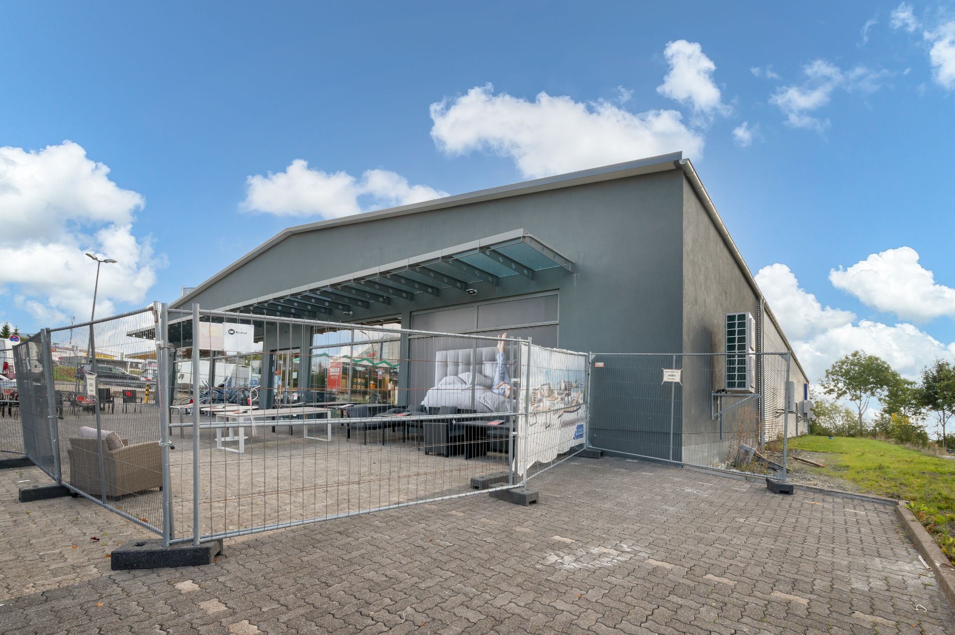 Grau gestrichenes Gebäude mit Glasvordach und Bauzaun auf gepflastertem Hof.