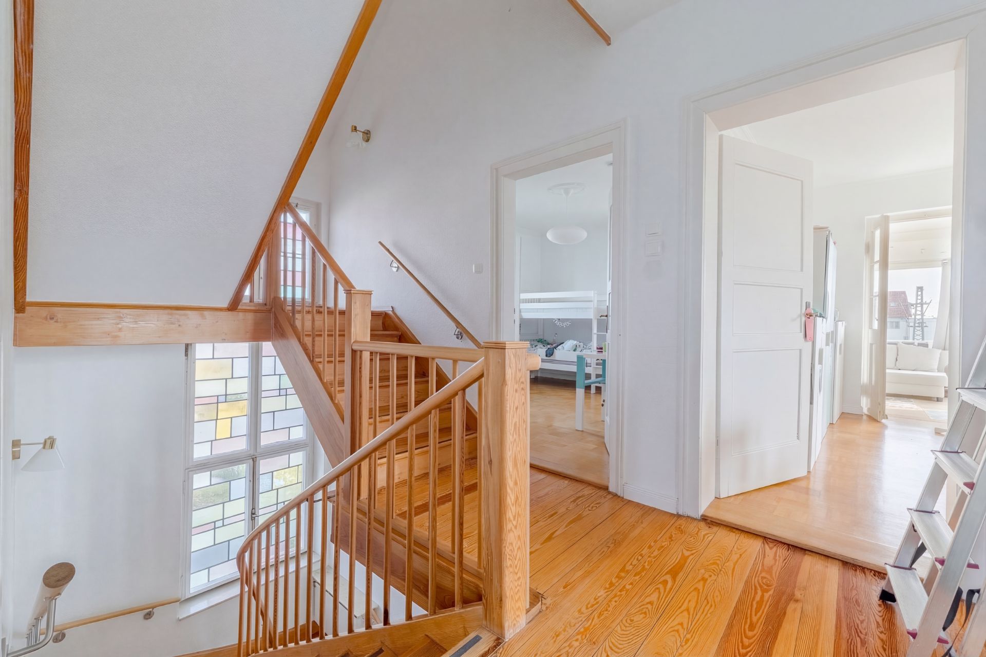 Heller Hausflur mit Holztreppe, zwei offenen Türen und großen Fenstern mit buntem Glas.