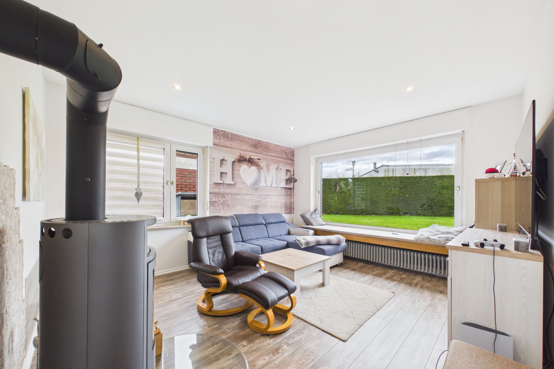 Helles Wohnzimmer mit Sofa, Sessel, Ofen, großem Fenster und Holzboden.
