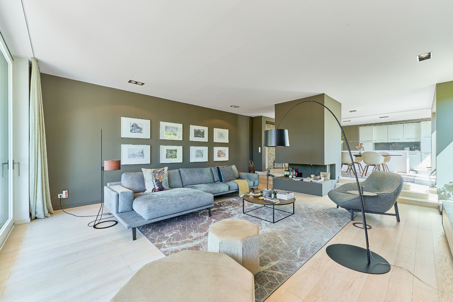 Helles Wohnzimmer mit grauem Sofa, Sessel, Teppich und Wandbildern, angrenzend an Ess- und Küchenbereich.