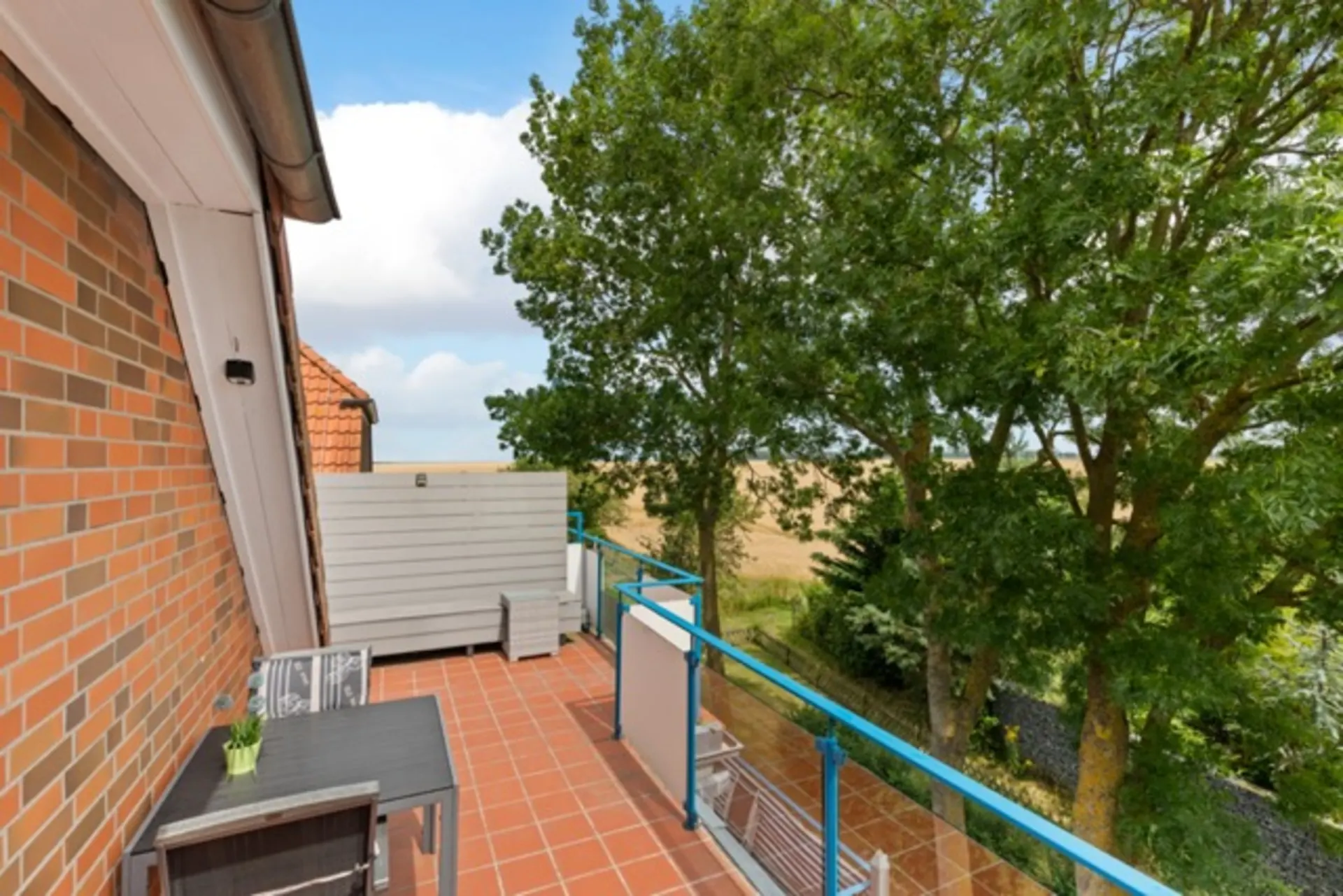 Balkon mit Terrakottafliesen, Tisch, zwei Stühlen und Blick auf Bäume und ein Feld.