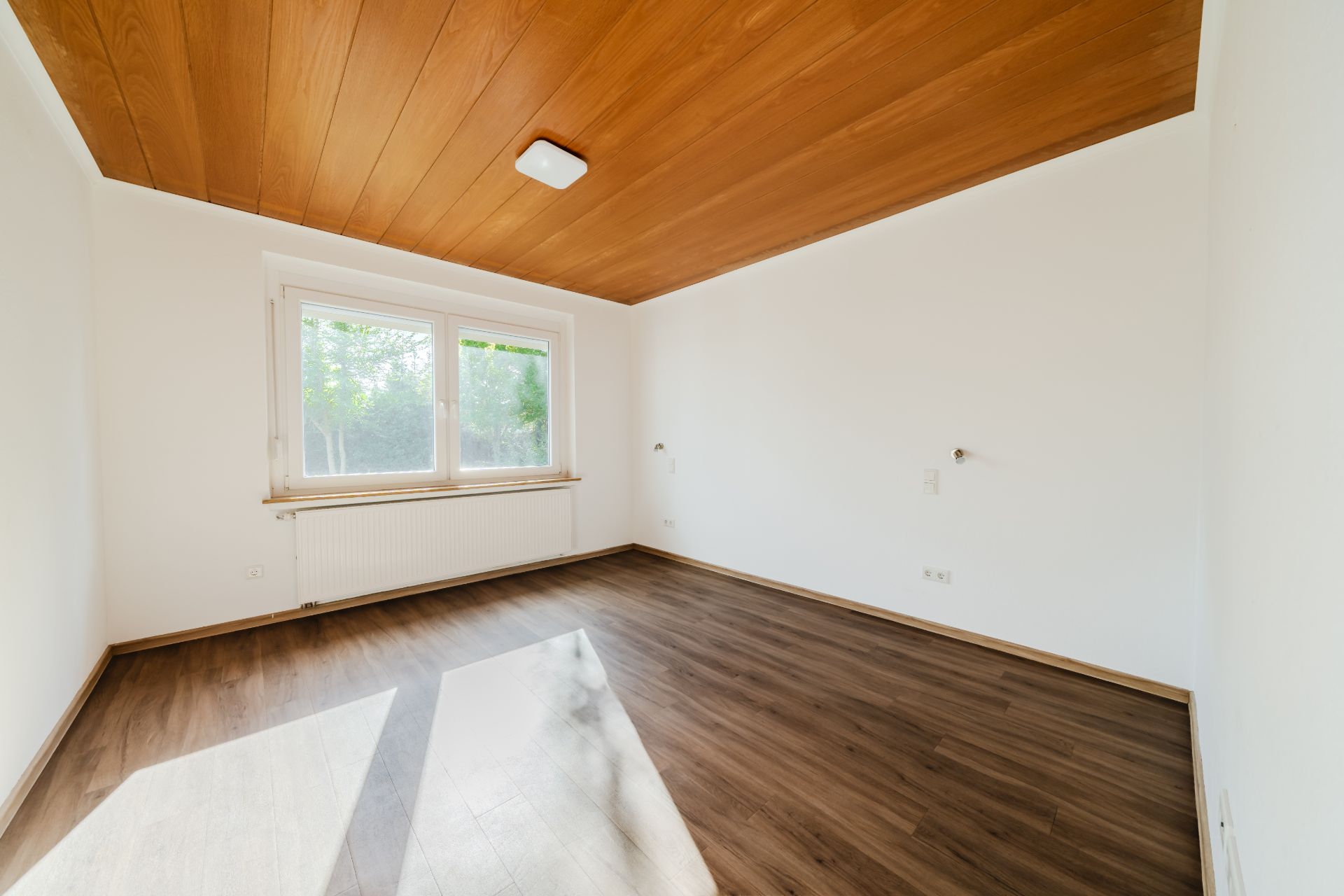 Helles, leeres Zimmer mit Holzdecke, großem Fenster und dunklem Boden.