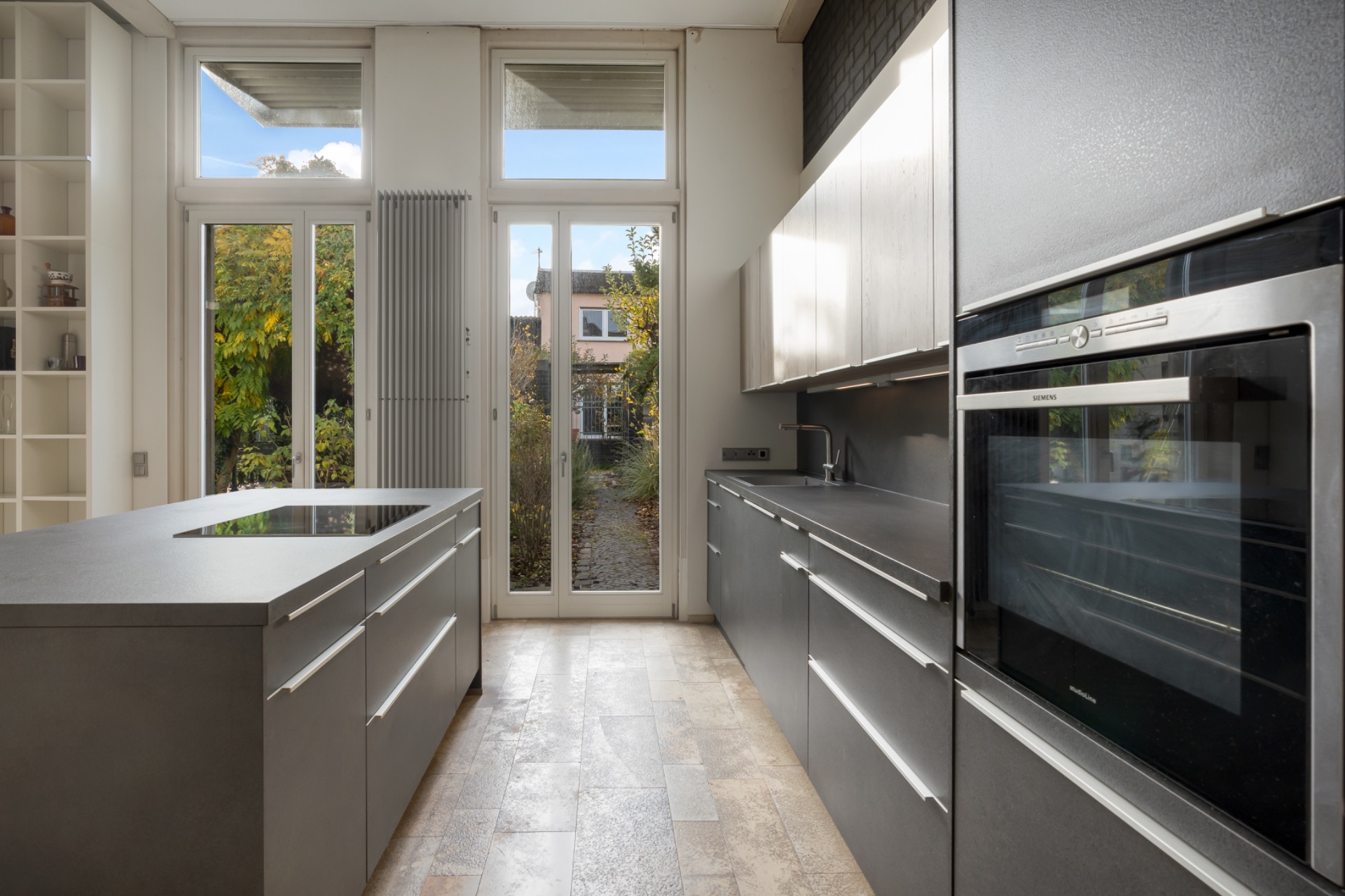 Moderne Küche mit Kochinsel, grauen Schränken und großen Fenstern zum Garten.