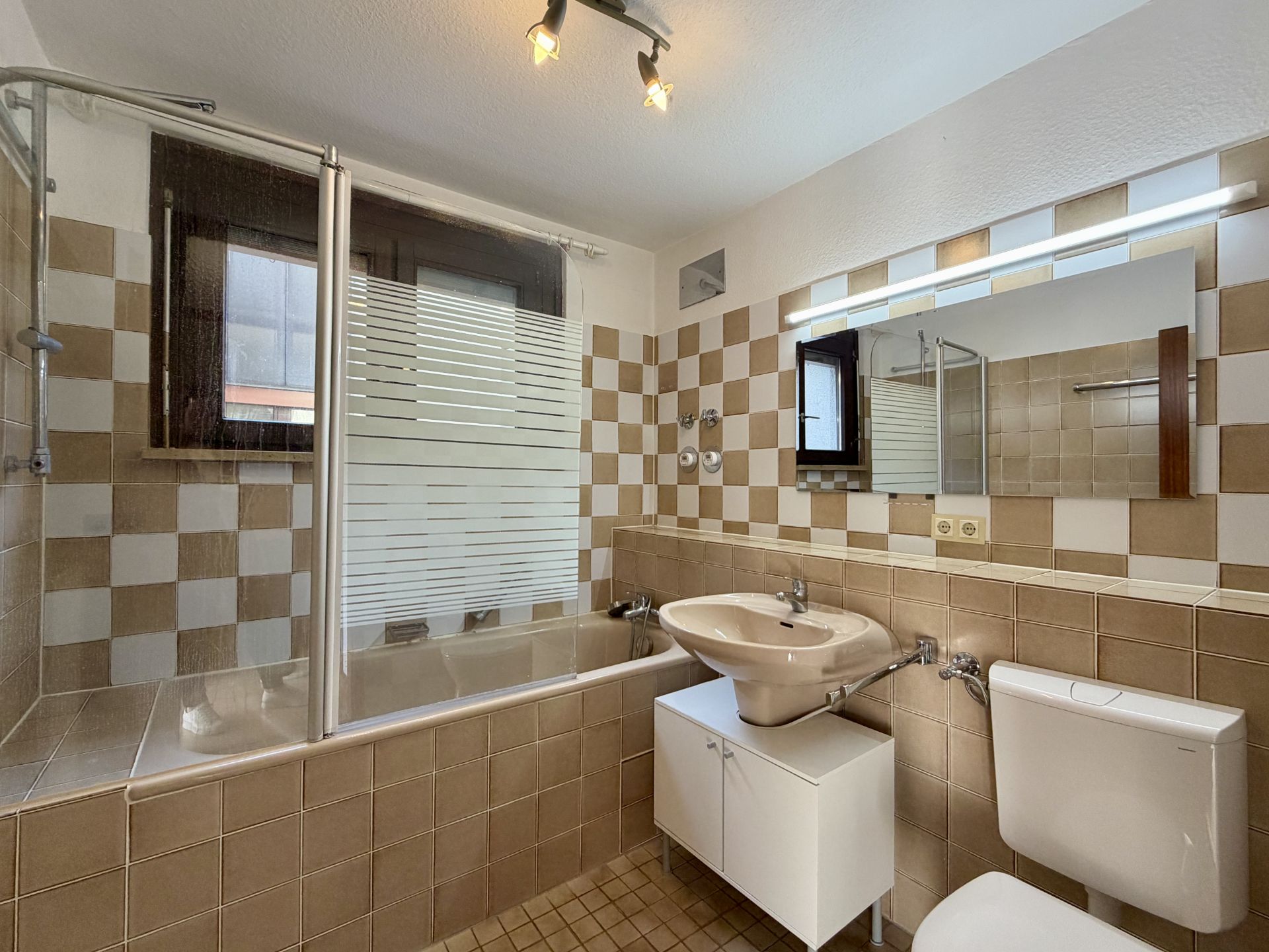 Badezimmer mit braun-weißen Fliesen, Badewanne, Waschbecken, Spiegel und Fenster.