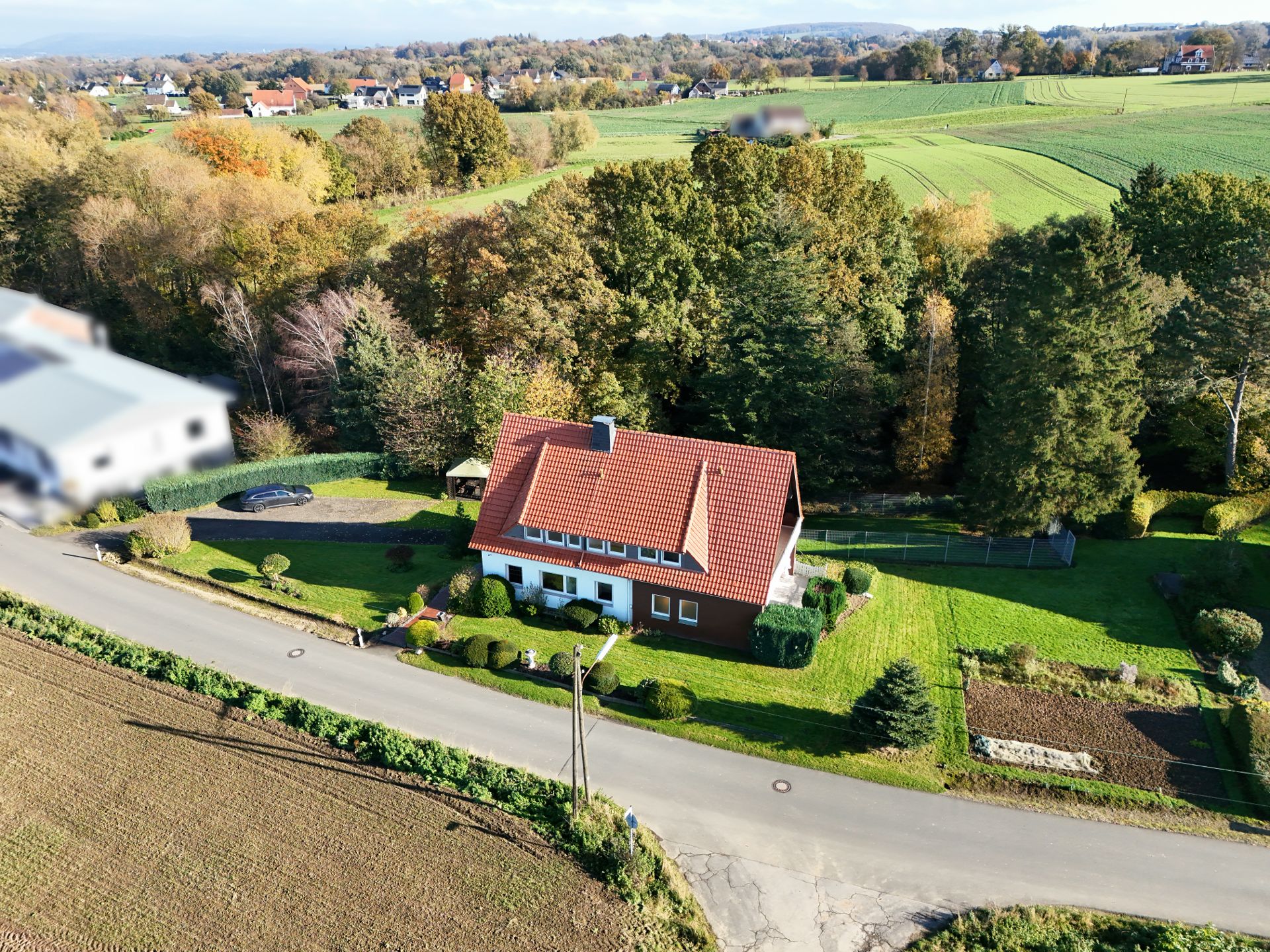 Luftaufnahme eines Einfamilienhauses mit rotem Dach und großem Garten an einer Landstraße.