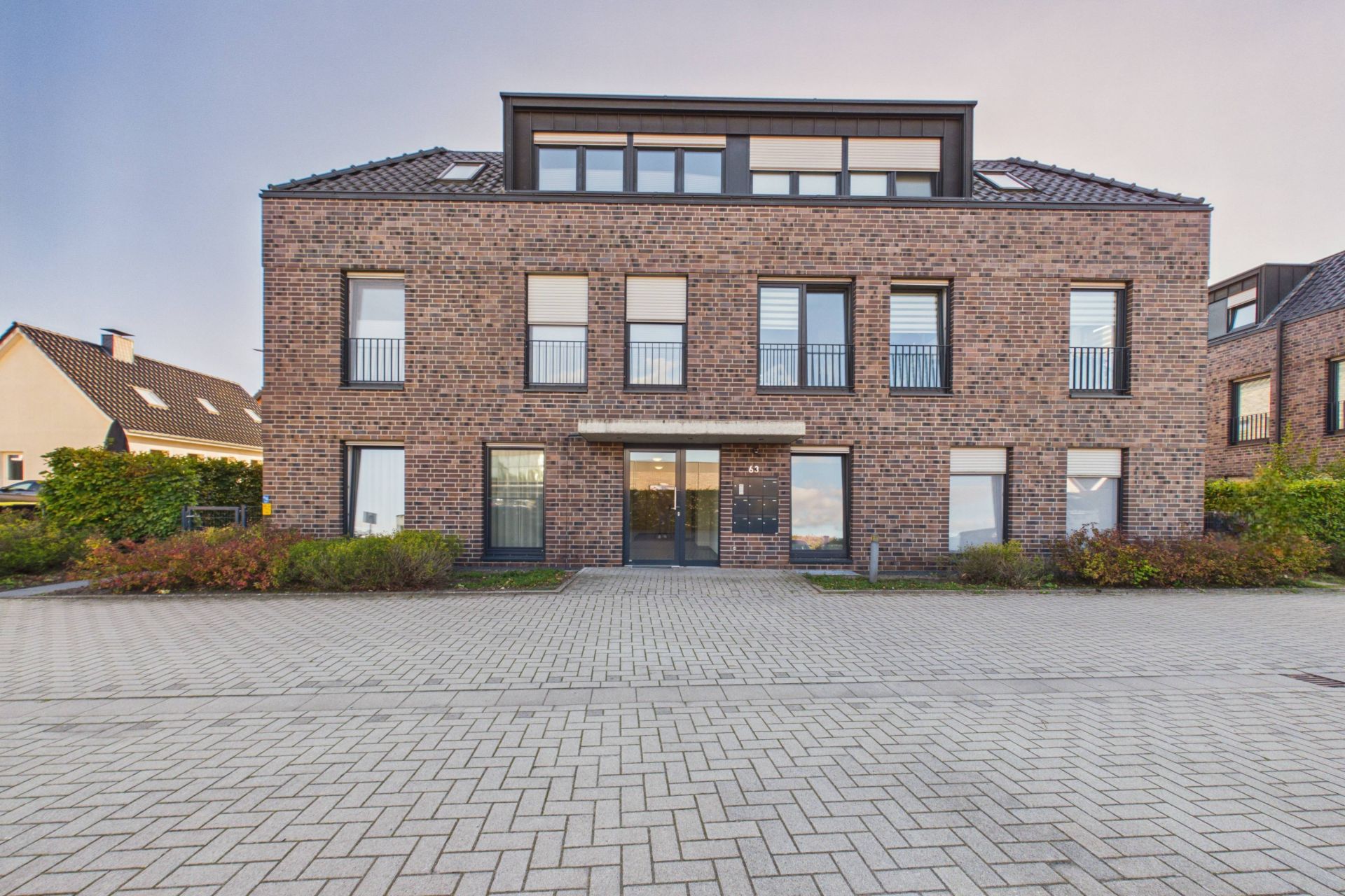 Modernes Wohngebäude mit Backsteinfassade und mehreren Fenstern, davor gepflasterter Hof.