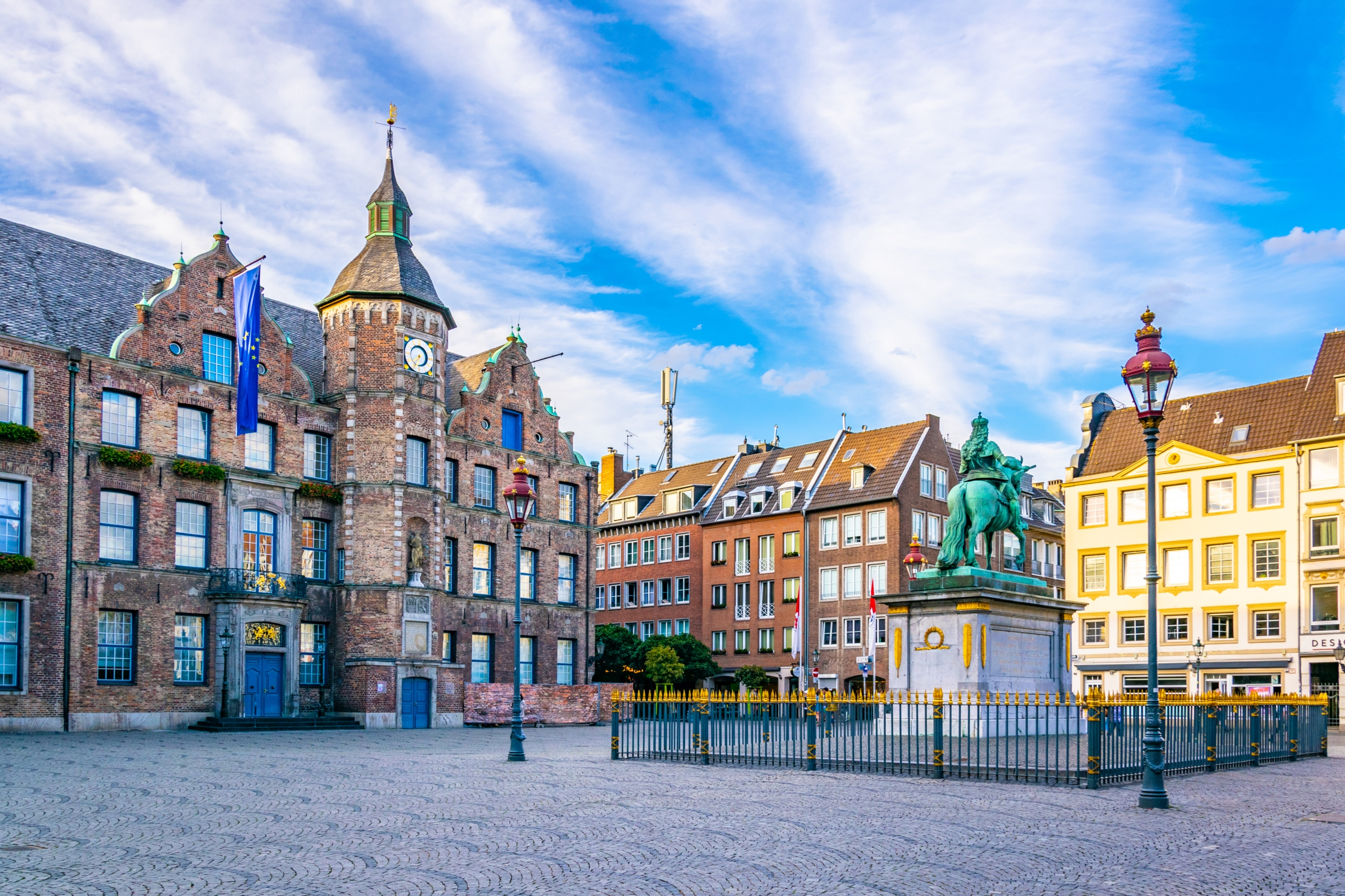 Historisches Gebäude mit Uhrturm und Reiterstatue auf einem offenen Platz, umgeben von Wohnhäusern.