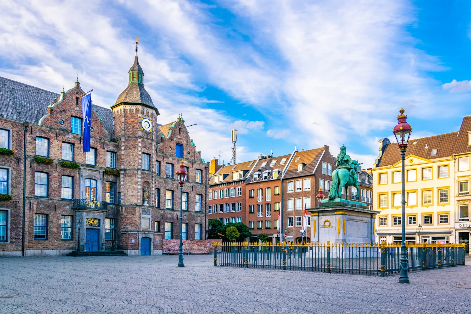 Historisches Gebäude mit Uhrturm und Reiterstatue auf einem offenen Platz, umgeben von Wohnhäusern.