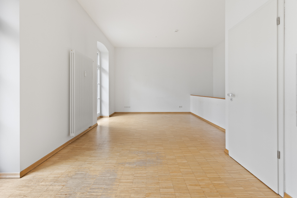 Heller, leerer Raum mit Holzboden, weißen Wänden und großem Fenster.