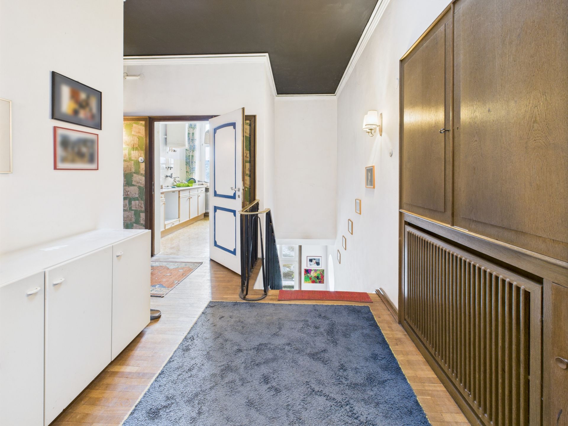 Flur mit Teppichboden, weißen und Holzmöbeln, Treppe und geöffneter Tür zur Küche.