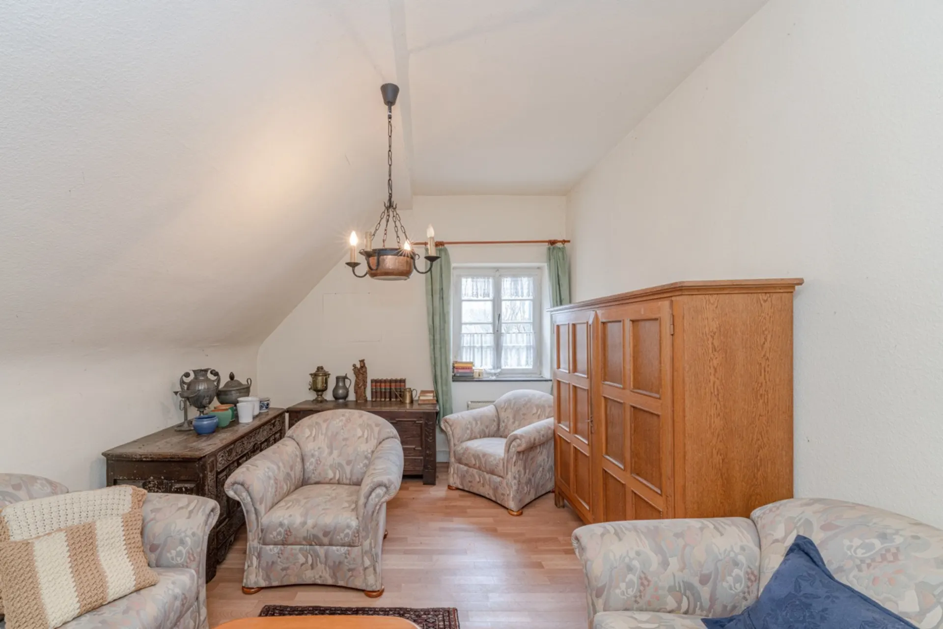 Gemütliches Wohnzimmer mit Sesseln, Holzschrank und kleinem Fenster mit Gardinen.